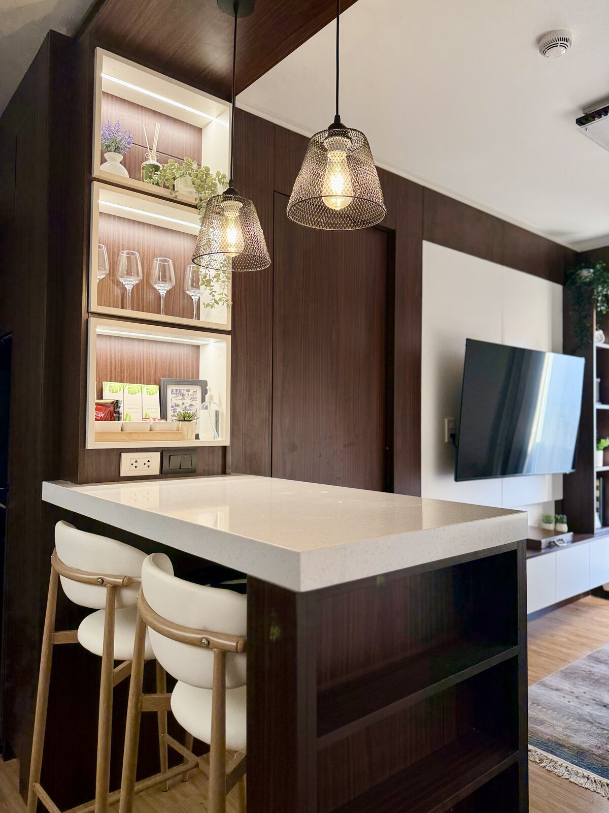 Dining counter with two stools under glass pendants, beside a lit niche of wine glasses.